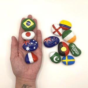 May include: A collection of painted rocks featuring flags from various countries, including Brazil, Japan, Australia, France, New Zealand, Ireland, Saudi Arabia, Sweden, Pakistan, and Aboriginal Australia.
