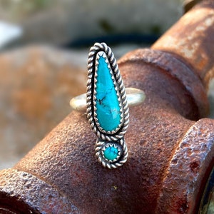 Op de afbeelding: Een zilveren ring met een turquoise steen in een touwontwerp. De ring ligt op een roestige pijp.