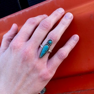 Op de afbeelding: Een zilveren ring met een turquoise steen in een puntige vorm. De ring wordt gedragen aan de middelvinger van een hand.