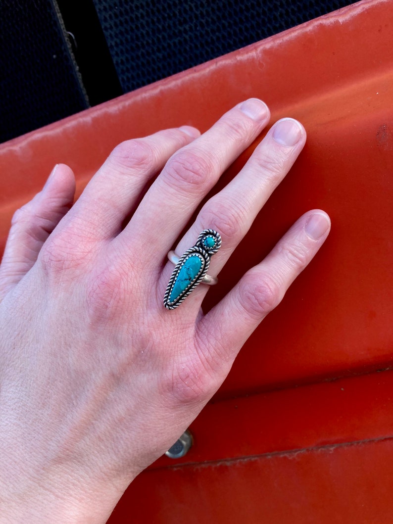 Op de afbeelding: Een zilveren ring met een turquoise steen in een puntige vorm. De ring wordt gedragen aan de middelvinger van een hand.