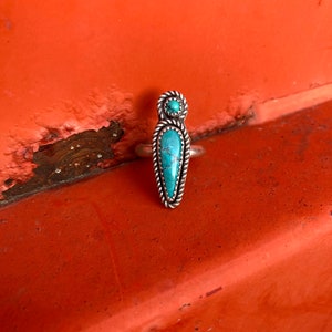 Op de afbeelding: Een zilveren ring met twee turquoise stenen. De grotere steen is lang, puntig ovaal en de kleinere steen is rond. De ring ligt op een rode achtergrond.