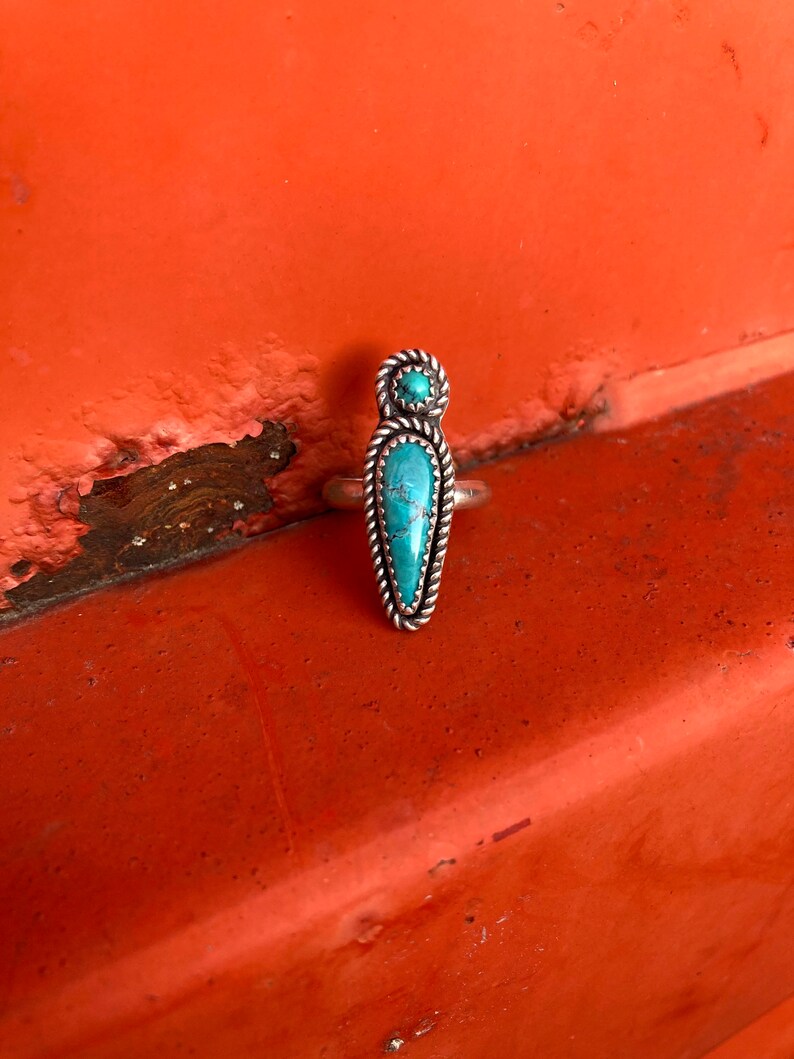 Op de afbeelding: Een zilveren ring met twee turquoise stenen. De grotere steen is lang, puntig ovaal en de kleinere steen is rond. De ring ligt op een rode achtergrond.