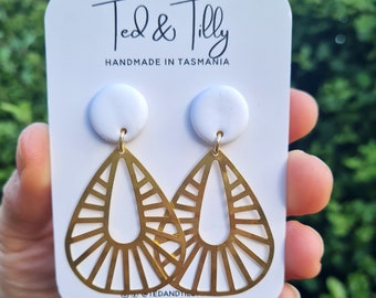Op de afbeelding: Een paar gouden oorbellen met een geometrisch ontwerp en witte keramische toppen. De oorbellen worden getoond op een witte kaart met de tekst "Ted & Tilly Handmade in Tasmania".