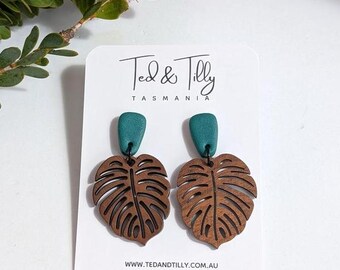 Op de afbeelding: Turquoise en bruine houten oorbellen met een gesneden monstera-bladontwerp. De oorbellen worden gepresenteerd op een witte kaart met de tekst "Ted & Tilly Tasmania" en website-informatie.
