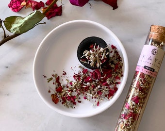 Könnte beinhalten: Ein kleines Glasröhrchen gefüllt mit getrockneten Kräutern und Rosenblüten mit der Aufschrift "KAYA BOTANICALS VENUS Love Smudge Blend". Eine weiße Keramikschale mit einer schwarzen Kohletablette und verstreuten Kräutern und Rosenblüten.