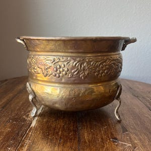 May include: A gold-toned metal planter with a grapevine design and three legs. The planter is on a wooden surface.