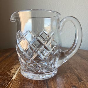 Może przedstawiać: Przezroczysty szklany dzbanek z diamentowym wzorem na dolnej części i zakrzywionym uchwytem. Dzbanek ma dzióbek ułatwiający nalewanie i stoi na okrągłej podstawie. Dzbanek jest wykonany z przezroczystego szkła.