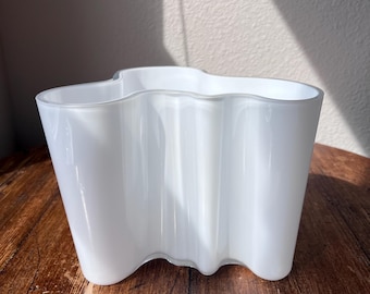 May include: A white glass vase with a wavy, organic design. The vase has a wide opening and a curved base. The smooth, opaque surface reflects light, creating highlights and shadows. The vase is likely for decorative purposes.