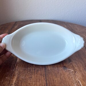 Może przedstawiać: Owalne, kremowe naczynie do serwowania z dekoracyjnymi uchwytami. Naczynie wykonane jest z ceramiki lub porcelany i stoi na drewnianej powierzchni. Uchwyty mają kwiatowy wzór. Naczynie jest puste.