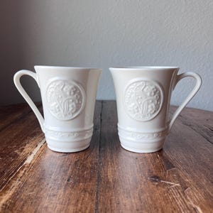 May include: Two white ceramic mugs with a decorative embossed design. Each mug features a circular emblem with floral and scroll details, and a curved handle. The mugs are set on a wooden surface.