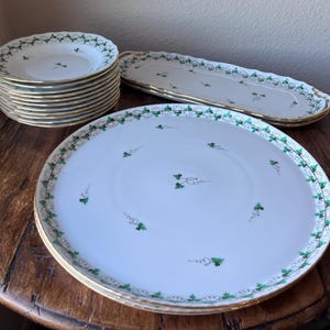 May include: A collection of white porcelain plates with a gold rim and green floral design. The set includes dinner plates, salad plates, and a serving platter. The plates are stacked on a wooden table.