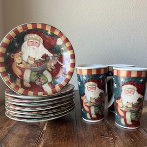 May include: A set of four Santa Claus themed mugs and a stack of six matching dinner plates. The mugs and plates feature a red and green colour scheme with a festive Christmas design. The mugs are white with a red and green Santa Claus design. The plates are white with a red and green Santa Claus design and a red and green border.