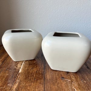 Puede incluir: Dos macetas cuadradas de cerámica de color blanco roto con bordes redondeados. Las macetas están vacías y se encuentran sobre una superficie de madera.