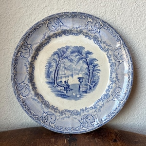 May include: A vintage blue and white porcelain plate with a decorative border and a central image of a landscape with trees, a bridge, and a building.