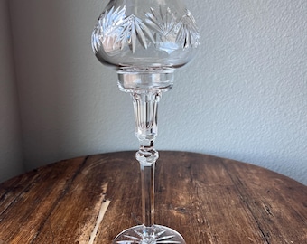May include: A clear crystal candle holder with a round base and a tall, slender stem. The top features a rounded, bell-shaped shade with etched leaf designs. The candle holder is displayed on a wooden surface.