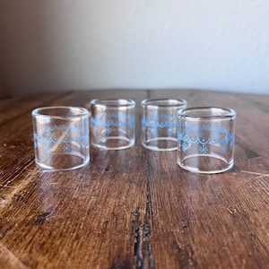 Puede incluir: Cuatro vasos de chupito de vidrio transparente con un patrón azul de copos de nieve y ondas alrededor del borde. Los vasos son cilíndricos y están sobre una superficie de madera.