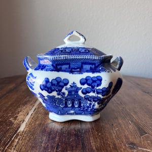 May include: A blue and white ceramic sugar bowl with a lid. The bowl features a classic Willow pattern design with a pagoda, trees, and a bridge. The bowl has two small handles and a decorative handle on the lid. The bowl is sitting on a wooden surface.