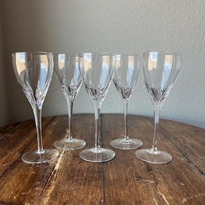 May include: Five clear crystal wine glasses with etched designs. The glasses have long stems and round bases, arranged on a rustic wooden table. The glasses are empty and reflect the light.