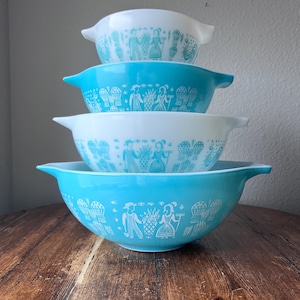 以下が含まれることがあります： 青と白の色合いの、4つのヴィンテージPyrexミキシングボウルのスタック。各ボウルは、人物と花の要素の白い様式化されたデザインが特徴です。ボウルは木製の表面に配置されています。