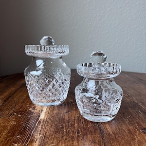 Könnte beinhalten: Zwei kleine, klare Glasbehälter mit Diamantschliffmustern und klaren, runden Deckeln. Die Gläser haben einen bauchigen Körper und einen dekorativen Rand. Auf einer dunklen Holzoberfläche.