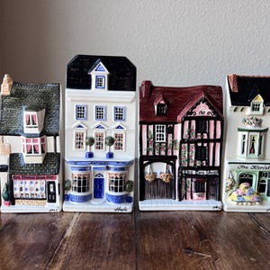 May include: Four ceramic miniature buildings with detailed facades. The buildings include a shop, an antique store, a tea shop, and a florist. Each building has a unique design, with varying roof styles, window arrangements, and decorative elements. The buildings are arranged on a wooden surface.