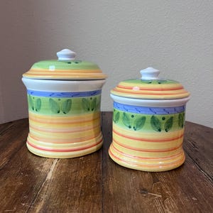 May include: Two ceramic canisters with lids. The canisters are cylindrical with a colourful design of horizontal stripes in yellow, orange, and red. The lids are green with a white knob. Blue and green leaf designs are painted on the canisters.