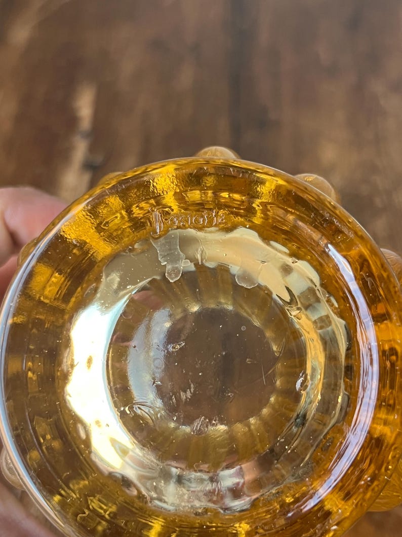 May include: The bottom of a clear amber glass vase with a textured surface. The vase has a round base and a wide opening. The text "131012" is visible on the bottom of the vase.