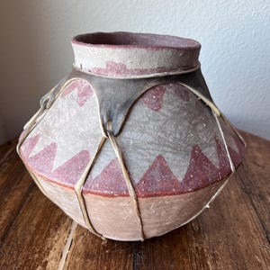 Könnte beinhalten: Ein handgefertigtes, rundes Töpfergefäß mit einer weiten Öffnung. Der Topf hat einen hellbraunen Boden, einen grauen Mittelteil mit roten Dreiecksmustern und einen grauen Rand. Natürliches Bindfaden sichert den Mittelteil. Das Gefäß wird wahrscheinlich zur Aufbewahrung oder Dekoration verwendet.