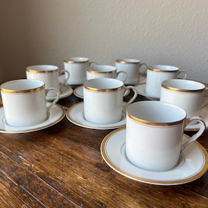 Puede incluir: Conjunto de diez tazas de café de porcelana blanca con ribete dorado. Cada taza tiene un platillo. Las tazas y los platillos están dispuestos en círculo sobre una mesa de madera.