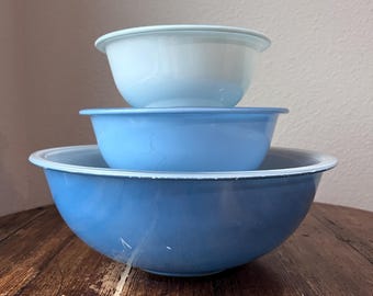 May include: Three blue enamel mixing bowls stacked on a wooden surface. The bowls vary in shades of blue, with white rims. The largest bowl is a darker blue, the middle bowl is a medium blue, and the top bowl is a light blue.