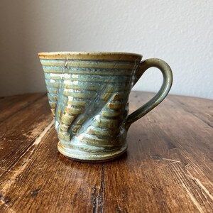 Könnte beinhalten: Eine handgefertigte Keramiktasse mit grüner Glasur und braunen Akzenten. Die Tasse hat ein strukturiertes, wellenförmiges Design und einen abgerundeten Griff.