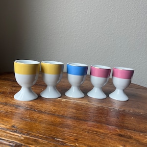 以下が含まれることがあります： 色のついた帯が付いた白い陶器製の卵立て6個セット。卵立ての上部には黄色、青、ピンクの帯が付いています。卵立ては木製の表面に一列に並べてあります。