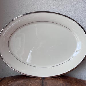 May include: An oval serving platter with a cream-colored surface and a silver-colored rim. A thin silver line runs along the inner edge of the platter. The platter is set on a wooden surface.