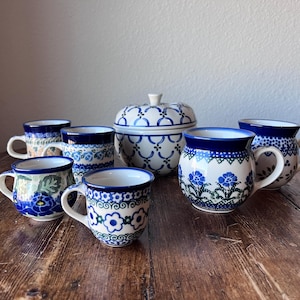 May include: A collection of ceramic mugs and a lidded bowl with blue floral patterns and dark blue rims. The mugs vary in size and design, with white backgrounds and intricate blue flower motifs. The bowl has a matching design.