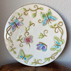 May include: A white ceramic plate with a gold rim and a design of butterflies in various colors, including purple, blue, green, and pink. The butterflies are surrounded by gold vines and leaves.