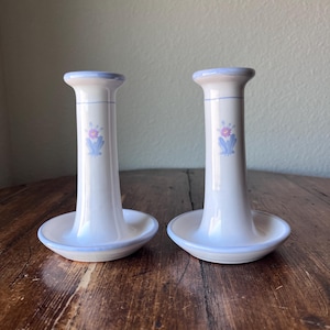 May include: Two white ceramic candle holders with a light blue rim and floral design. The candle holders have a round base and a tall, cylindrical body. The floral design features purple and blue flowers.