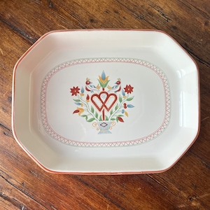 Puede incluir: Un plato de servir de cerámica blanca con un diseño floral rojo, verde, amarillo y azul con dos pájaros y un corazón. El plato tiene un borde festoneado y un borde rojo.