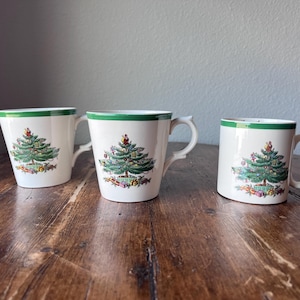 Puede incluir: Tres tazas de cerámica blanca con borde y asa verdes. Cada taza presenta un diseño detallado de árbol de Navidad con adornos y regalos. Las tazas están sobre una superficie de madera.