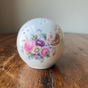 Puede incluir: Porta-potpourri de cerámica blanca con un diseño floral con flores rosas, moradas y amarillas y una mariposa. El porta-potpourri tiene pequeños agujeros alrededor de la parte superior.