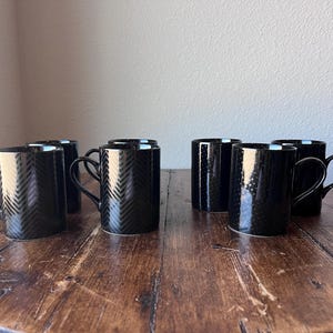 May include: A set of six black ceramic mugs with a textured chevron pattern. The mugs have a glossy finish and curved handles. They are arranged on a dark wooden surface.