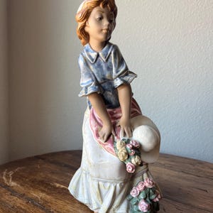 Puede incluir: Una figurita de porcelana de una niña que lleva un vestido azul y blanco y una faja rosa. Lleva un sombrero blanco y un ramo de rosas rosas y amarillas.