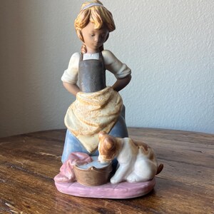 Puede incluir: Una figurita de porcelana de una niña con un vestido azul y un delantal marrón, sosteniendo un cubo mientras un pequeño perro blanco con manchas marrones bebe de él.