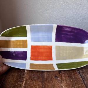 Könnte beinhalten: Eine rechteckige Servierplatte aus Keramik mit einem farbenfrohen geometrischen Design. Die Platte hat ein Rastermuster aus Quadraten in Grün-, Orange-, Lila-, Blau- und Beigetönen, getrennt durch weiße Linien. Die Platte wird von einer Hand gehalten.