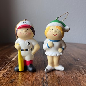 Puede incluir: Dos adornos navideños de cerámica que representan a una niña y un niño. La niña lleva un vestido blanco y un gorro de Papá Noel verde. El niño lleva un uniforme de béisbol con un gorro rojo y un bate amarillo. Ambos adornos tienen caras pintadas con mejillas sonrosadas.