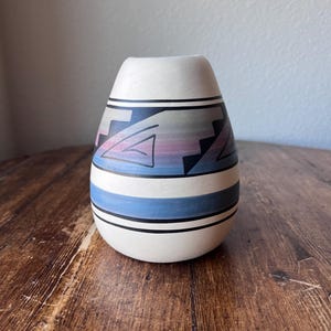 以下が含まれることがあります： 丸みを帯びたベースと細い首を持つ、クリーム色のセラミック花瓶。花瓶には、青、黒、ピンクの幾何学模様が施されています。水平の青いストライプと黒い線がベースを囲んでいます。花瓶は木製の表面に飾られています。