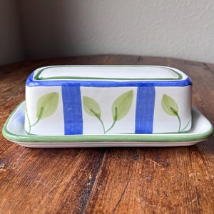 Könnte beinhalten: Eine rechteckige Butterdose aus Keramik mit weißem Unterteil und Deckel. Der Deckel ist mit vertikalen blauen Streifen und handgemalten grünen Blattmotiven verziert. Die Ränder des Deckels und der Basis sind mit einer grünen Linie umrandet.