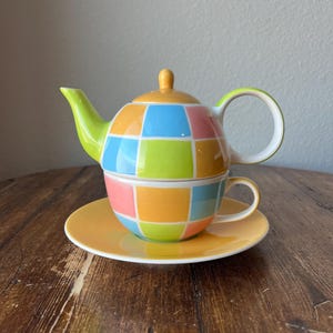 Könnte beinhalten: Ein farbenfrohes Teeset mit einer Teekanne und einer Tasse, beide mit einem geometrischen Muster aus Quadraten in Gelb, Grün, Blau und Rosa.