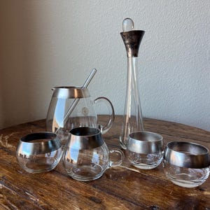 Może przedstawiać: Zestaw szklanych kubków z srebrnymi obwódkami, szklany dzbanek z srebrną obwódką i szklaną pałeczką do mieszania. Zestaw zawiera również szklaną karafkę ze srebrnym korkiem.