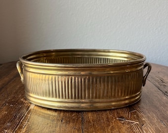 May include: Oval brass planter with two handles. The planter has a ribbed design and is made of shiny brass.