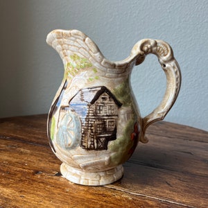 May include: A ceramic pitcher with a brown, green, and white painted design of a watermill and a house. The pitcher has a handle with a decorative design.